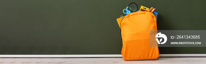 horizontal image of yellow backpack full of school stationery on desk near green chalkboard