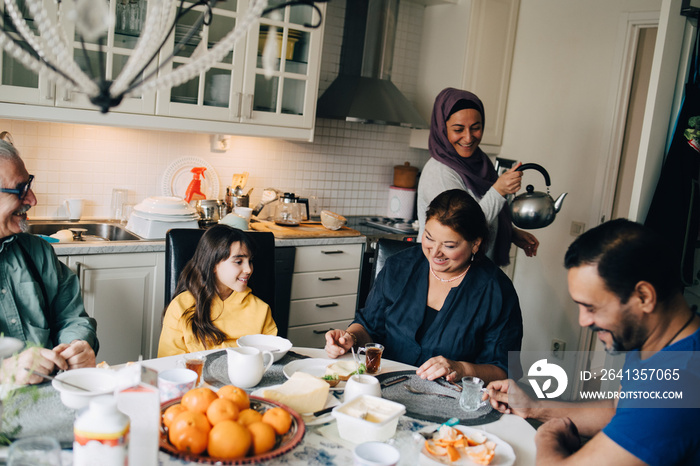 Mid adult woman serving tea to multi-generation family at dining table in kitchen