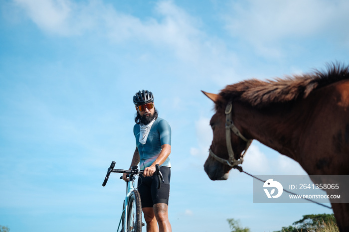 A young bearded cyclist is biking through a field with a horse