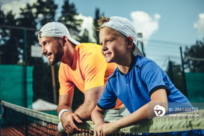 侧视积极的男孩和喜气洋洋的大胡子男性在打好网球后放松