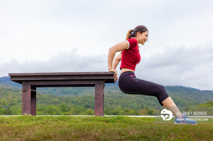 Beautiful young Asian woman doing tricep dips exercise outdoor at a park, cloudy sky and mountains b