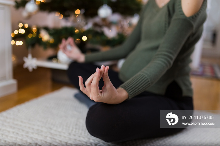 Close up of pregnant woman exercising yoga lotus position at home next to christmas tree.