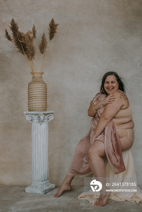 plus size metis woman holding pink fabric smiling at camera sitting next to roman column