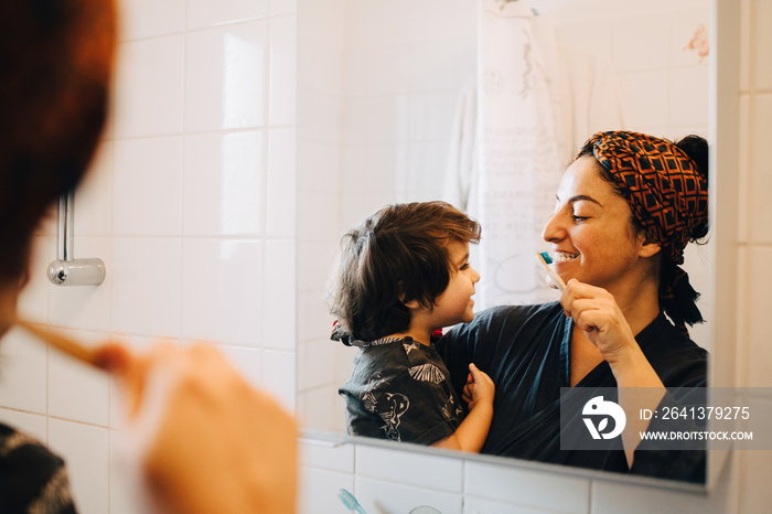 Mother carrying son while brushing teeth in front of mirror at home