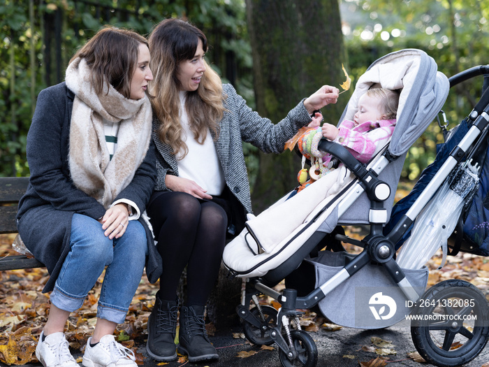 Lesbian couple sitting on bench next to a baby in stroller in park�