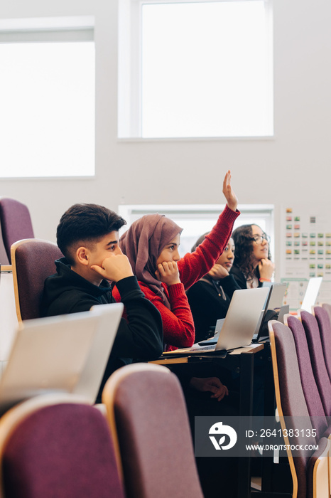 Teenage student in hijab raising hand while sitting with friends during lecture in classroom