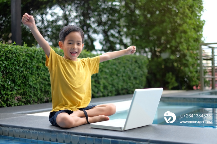 Happy Asian Young child using computer laptop with hand up at side of swimming pool