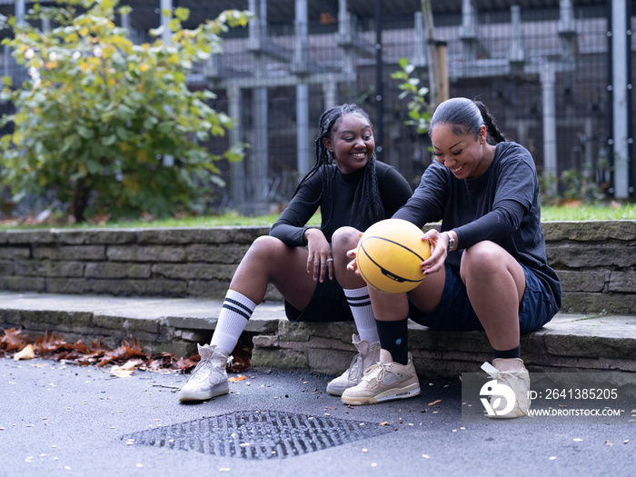 Two female friends sitting outdoors with basketball