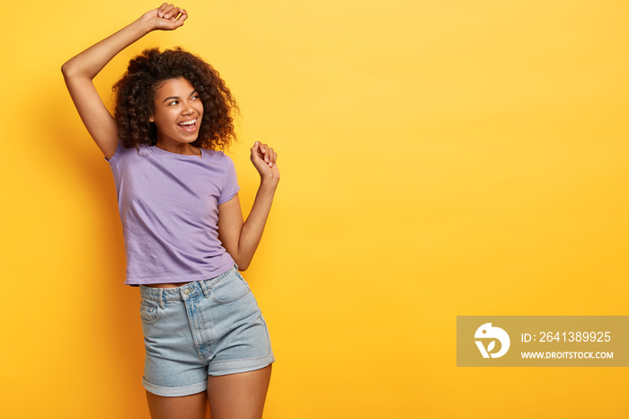 Good humored African American slim girl dances on yellow background, looks happily away, wears casua