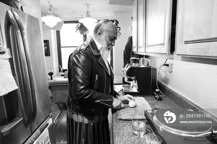 gray haired bearded man in leather jacket cuts a lemon a small kitchen