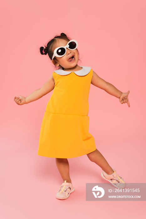 穿着黄色连衣裙，戴着墨镜，在粉色衣服上摆姿势的快乐亚洲幼童