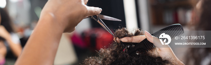 Cropped view of african american hairstylist holding comb and scissors near hair of client, banner