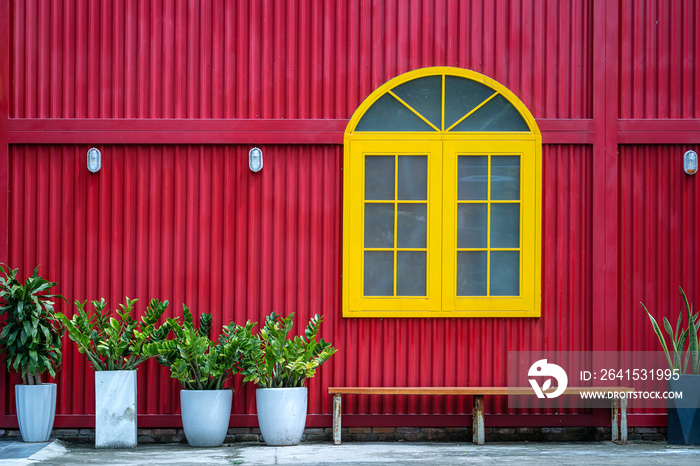 Yellow window, flowerpots with plants and bench on the background of a red metal wall on the street 