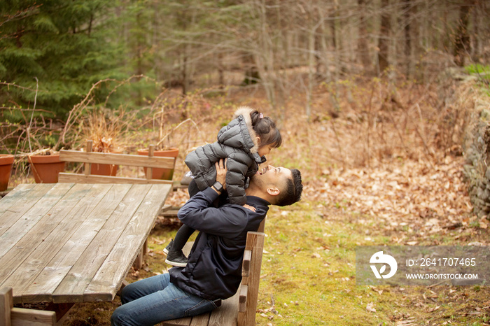 Side view of happy father picking up daughter while sitting on chair in forest