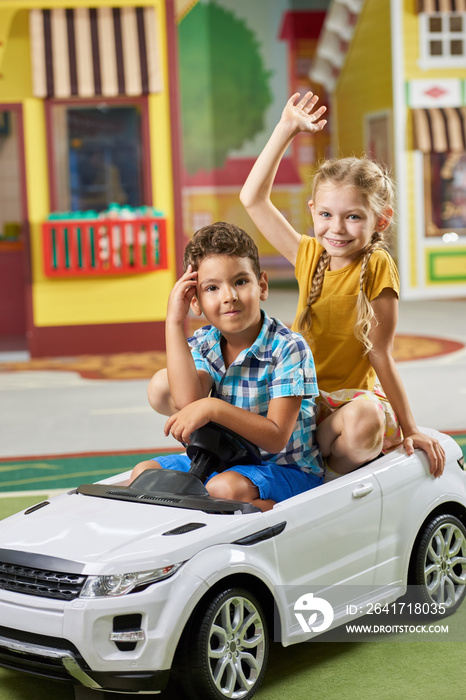 Happy kids sitting in toy car and looking at camera. Cute children having fun in play center. Kids l