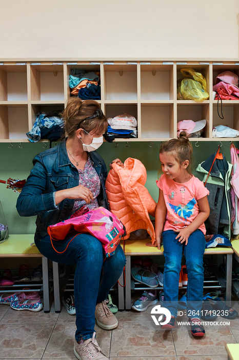 Child little girl changing clothes in changing room in nursery school with mothers help. Woman weari