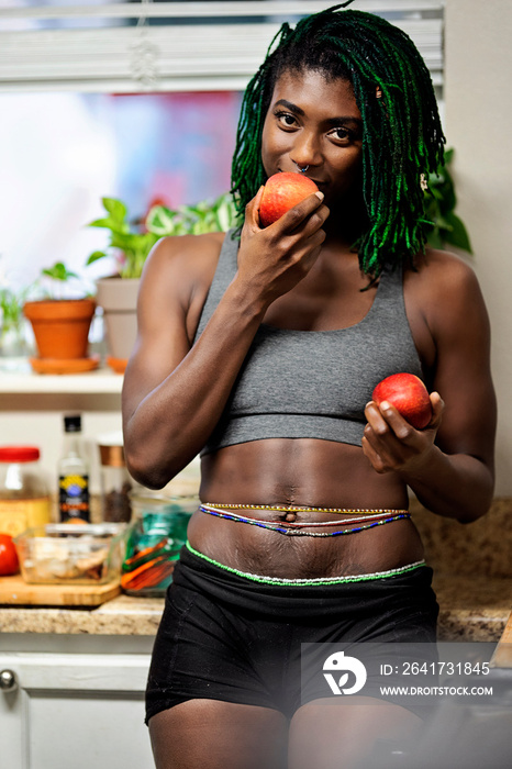 Black woman with green dreadlocks cleaning and eating fruit in her kitchen