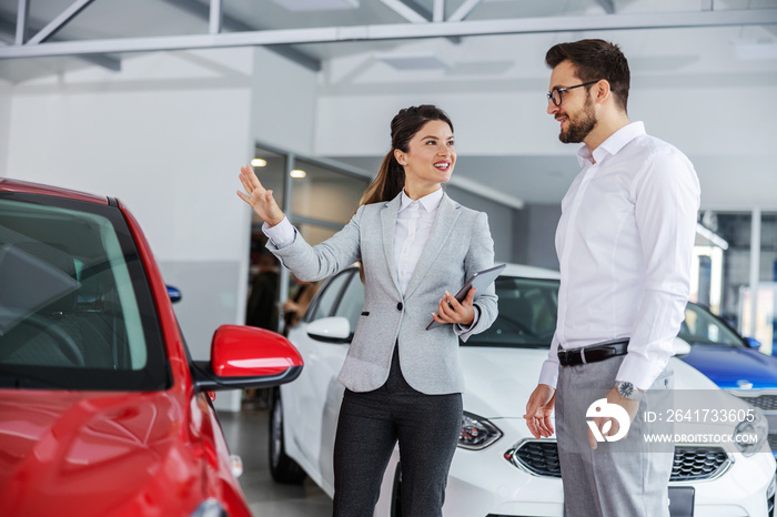 Smiling friendly female car seller with tablet in hands talking about car specifications to a man wh