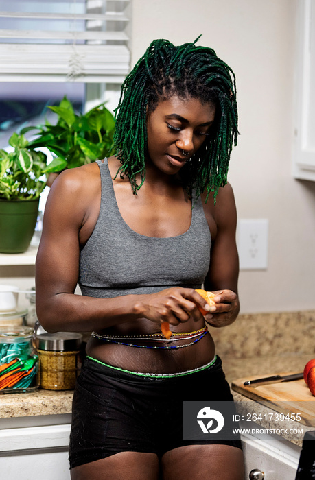 Black woman with green dreadlocks cleaning and eating fruit in her kitchen