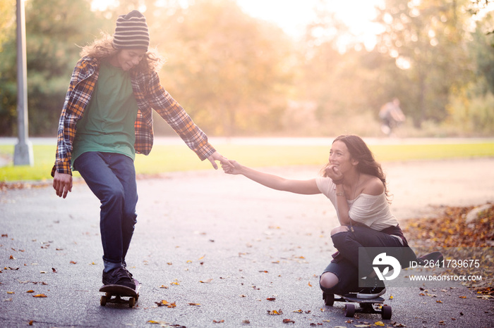 Couple skateboarding in park, holding hands, leaves on footpath