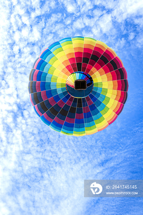 bunter Heißluftballon in Regenbogenfarben als Punkt von unten gegen den blauen Himmel mit weißen Wol