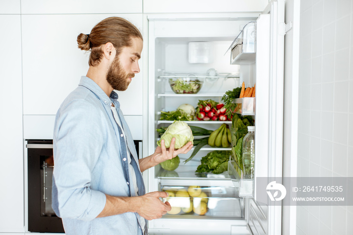 Young vegan man choosing what to cook, taking fresh vegetables from the refrigerator at home