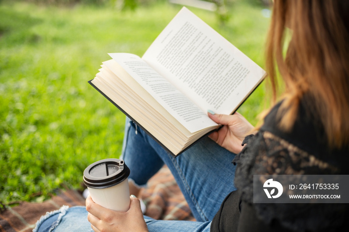 一个女人坐在公园的一棵树旁，手里拿着一本书和一个杯子，杯子里拿着热饮。哇
