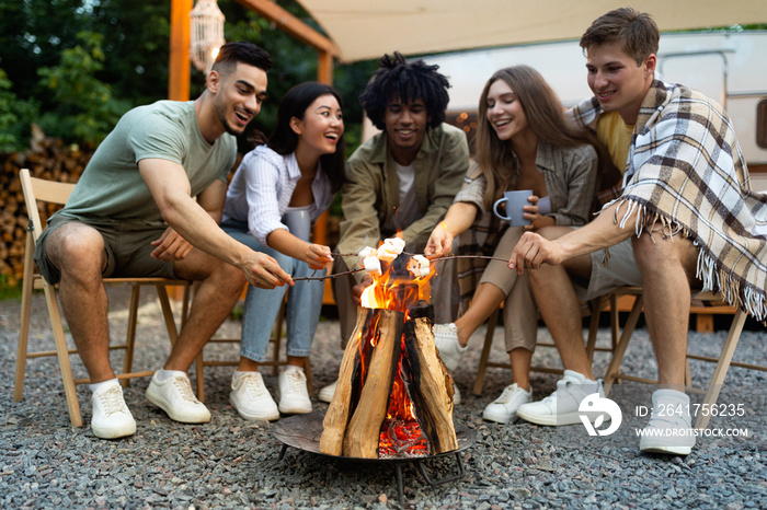 Young multiracial friends having outdoor picnic near RV, frying marshmallows on campfire, having fun