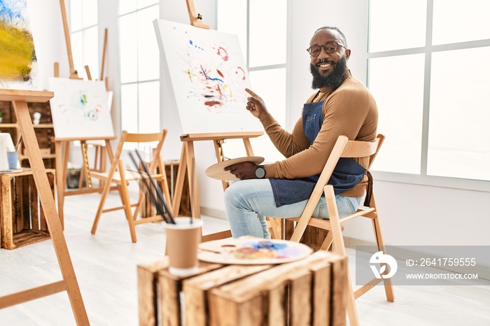 African american artist man painting on canvas at art studio smiling happy pointing with hand and fi