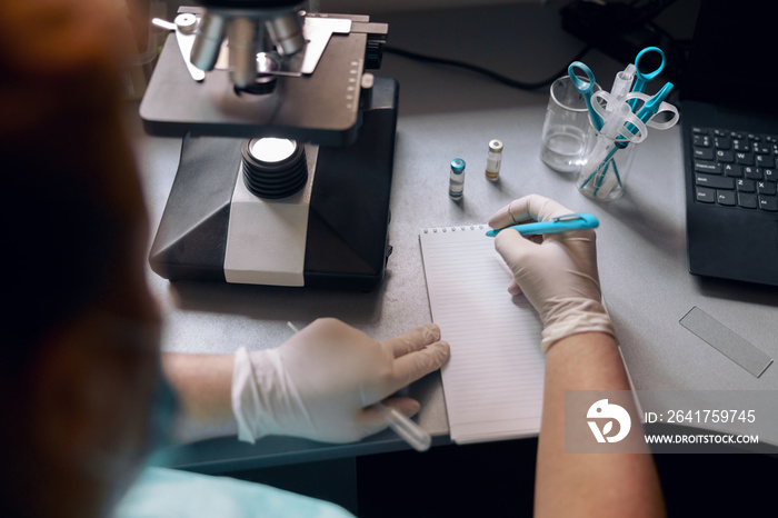 Lab tech in latex gloves writes marks in notebook near microscope at desk