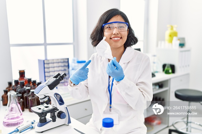 戴着安全口罩在科学家实验室工作的年轻西班牙妇女微笑着开心地指着哈