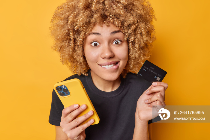 Indoor shot of curly woman bites lips looks surprisingly at camera holds smartphone and credit card 