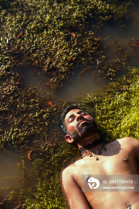 portrait of a dark skinned Indian man by a lake, shirtless, with make up, surrounded by nature