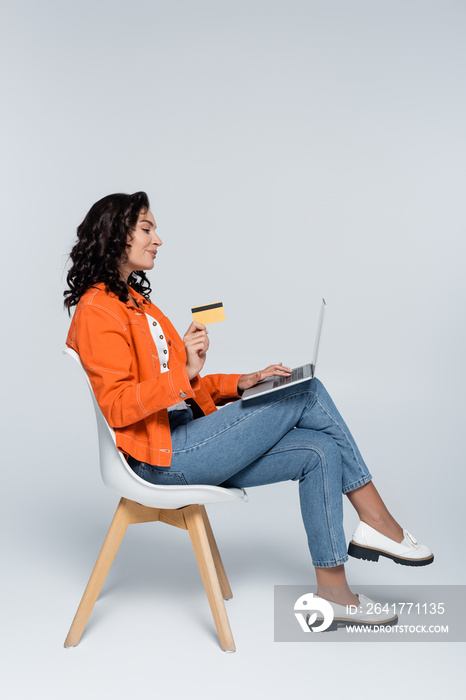 side view of young woman in orange jacket using laptop and holding credit card on grey.