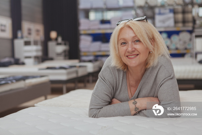 Happy relaxed mature woman smiling to the camera, lying on orthopedic mattress at furniture store. A