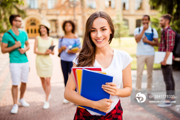 Focused shot of cheerful smart brunette caucasian girl is holding note books, smiling, standing near