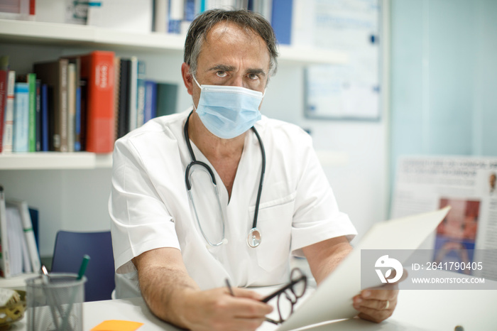 Medico cinquantenne in camice bianco e mascherina protettiva, seduto nella scrivania del suo ufficio