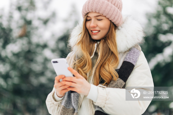 戴着针织帽，穿着保暖外套的女人，在冬季公园散步，用手机