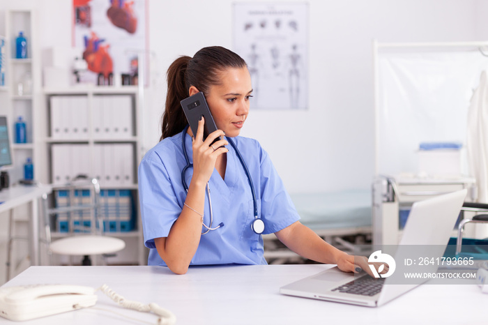 Medical practitioner using telephone and laptop in hospital office wearing blue uniform. Health care