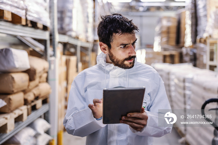 Young worker in sterile protective suit standing in warehouse with hand on hip and holding tablet wh