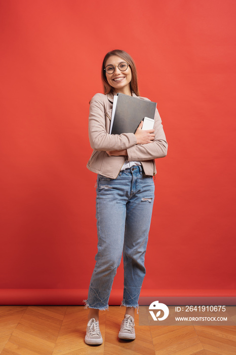 Young smiling student or intern in eyeglasses standing with a folder on red background.