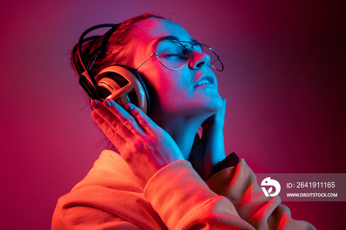 Fashion pretty woman with headphones listening to music over red neon background at studio.