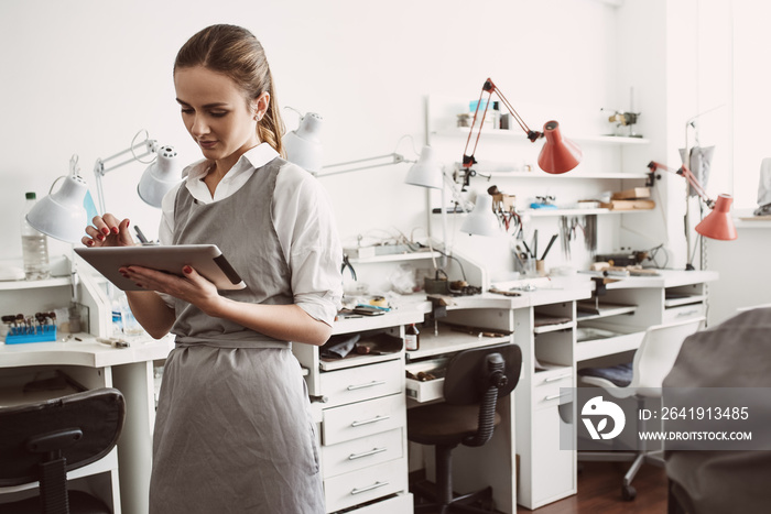 Online production. Close up portrait of young female jeweler in apron using CRM system on her digita