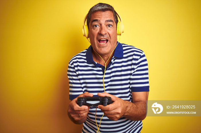 Middle age man playing video game using headphones over isolated yellow background scared in shock w