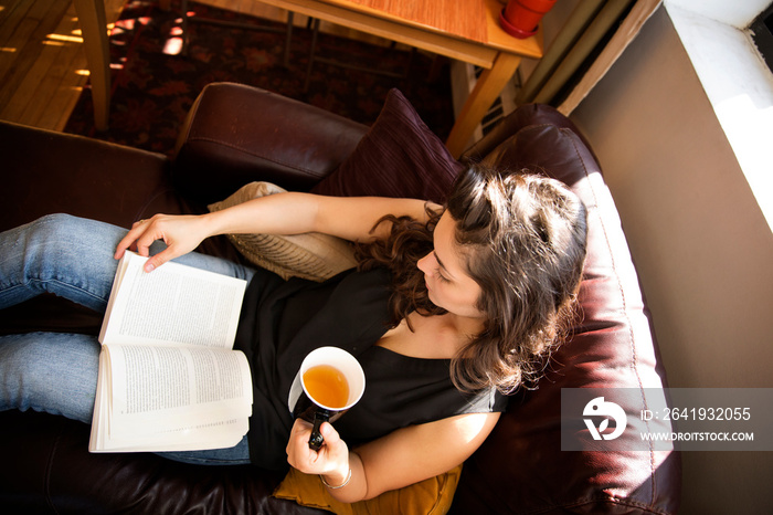 Young woman drinking tea and reading book on sofa
