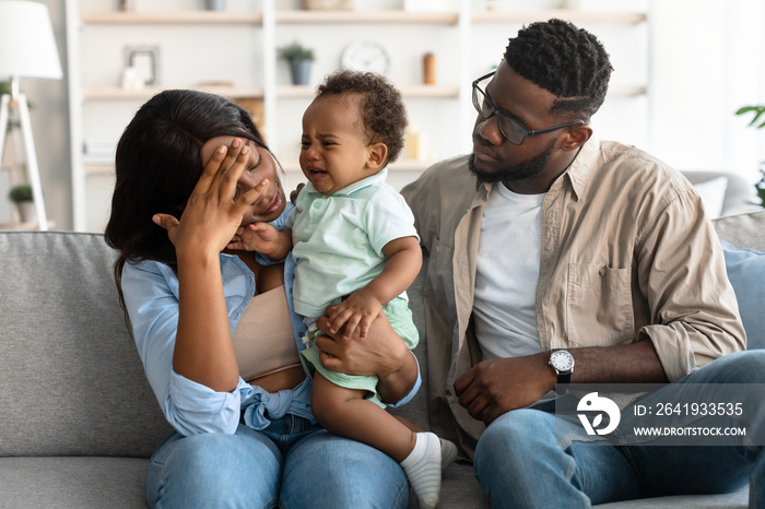 Tired African American parents sitting with crying kid on sofa