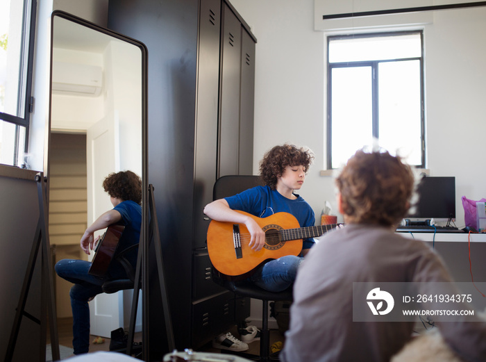 Rear view of boy looking at brother playing guitar while sitting on chair in bedroom