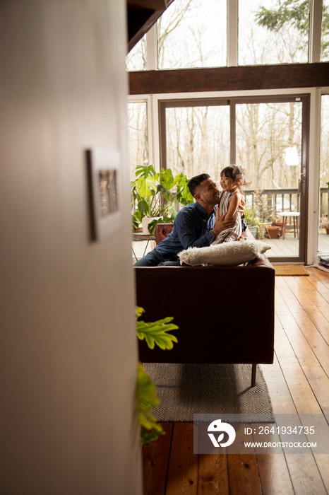 Side view of father with daughter relaxing on sofa against window at home seen through doorway