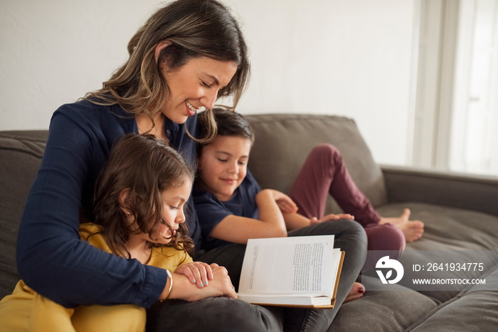 Smiling mother reading book for children while relaxing on sofa against wall at home