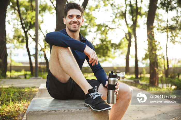 Portrait of beautiful athletic man sitting on bench and holding water bottle while doing workout in 